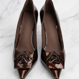 A pair of elegant dark chocolate brown patent leather pumps featuring a pointed toe and a sophisticated 3D rose floral decoration on the front, perfect for a quiet luxury office or evening look.