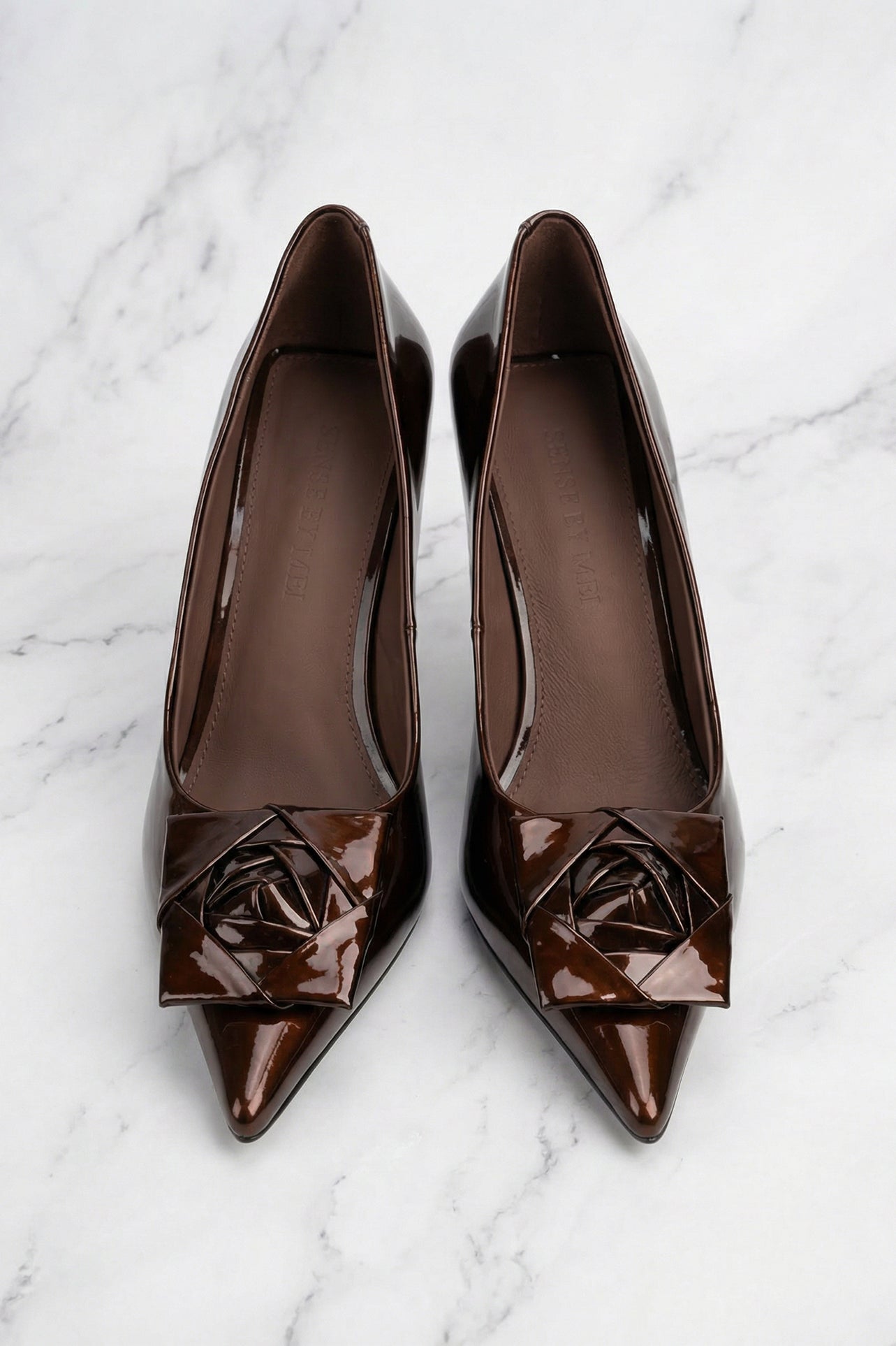 A pair of elegant dark chocolate brown patent leather pumps featuring a pointed toe and a sophisticated 3D rose floral decoration on the front, perfect for a quiet luxury office or evening look.