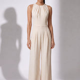 A style comparison of two elegant wide-leg jumpsuits; the left showcasing a sophisticated emerald green tailored pantsuit with a structured crop capelet sleeve, and the right featuring a minimalist cream white sleeveless jumpsuit with refined neck pleating details.