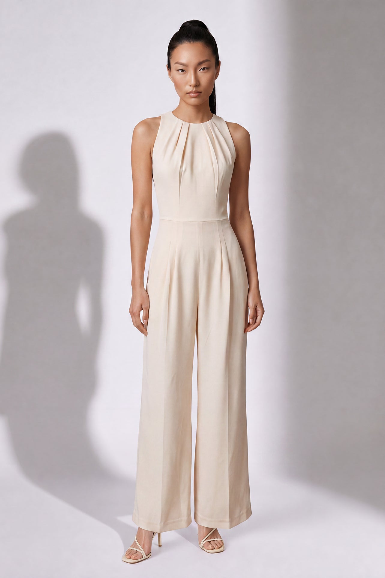 A style comparison of two elegant wide-leg jumpsuits; the left showcasing a sophisticated emerald green tailored pantsuit with a structured crop capelet sleeve, and the right featuring a minimalist cream white sleeveless jumpsuit with refined neck pleating details.