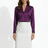 Two professional models wearing elegant V-neck silk satin blouses in shimmering silver and deep purple, featuring pleated front details and paired with white high-waisted trousers and a buttoned skirt.