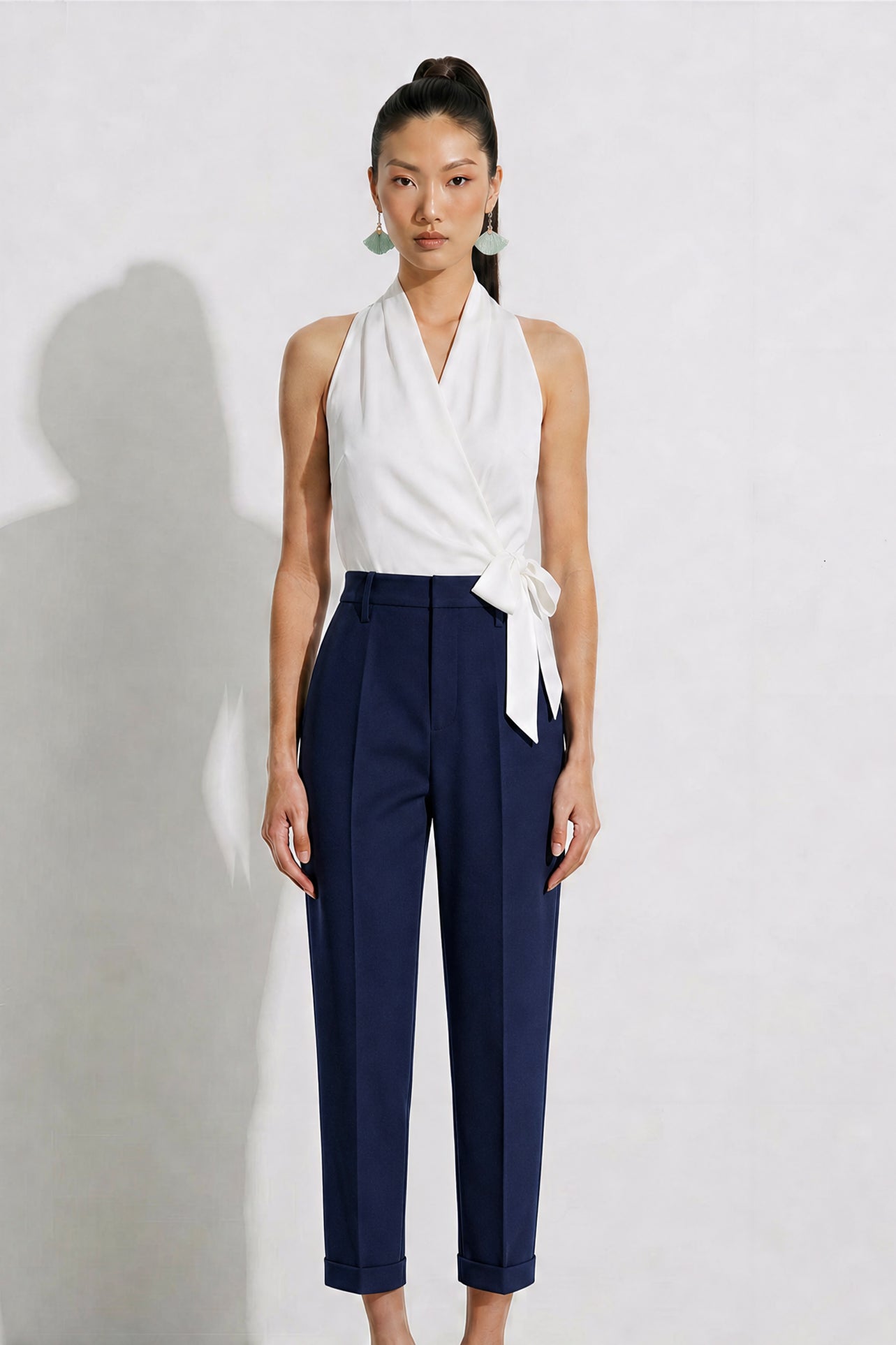 A model showcasing elegant navy blue high-waisted work trousers, featuring a unique curved overlay panel design at the waist, a slim golden metal bar buckle, pressed front creases, and chic cuffed hems, paired with a light blue button-up shirt.