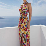 A beautiful professional model presenting an elegant tropical floral print halter maxi dress with a blue ocean background; featuring a sophisticated wrap tie-waist silhouette and vibrant multicolored botanical patterns for a chic summer resort look.