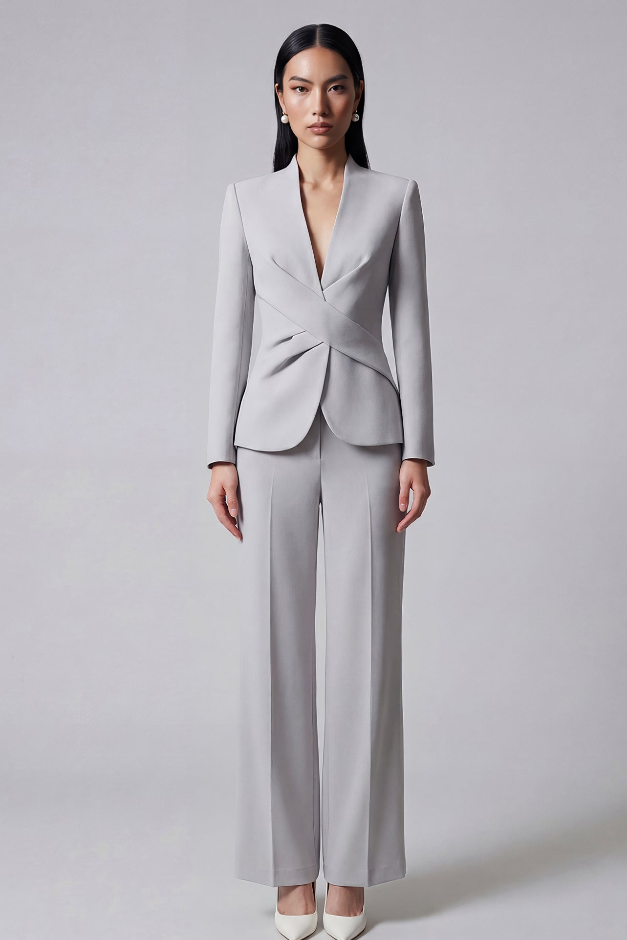 A professional woman wearing a sophisticated light grey tailored suit set, featuring a unique wrap-front blazer with criss-cross waist detailing and matching slim-fit trousers for a polished executive look.
