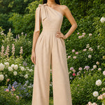 Two joyful women wearing elegant one-shoulder jumpsuits with large tie-bow shoulder straps in a sunny park; one jumps in a pastel pink wide-leg jumpsuit with yellow sneakers, while the other claps in a beige ankle-length jumpsuit