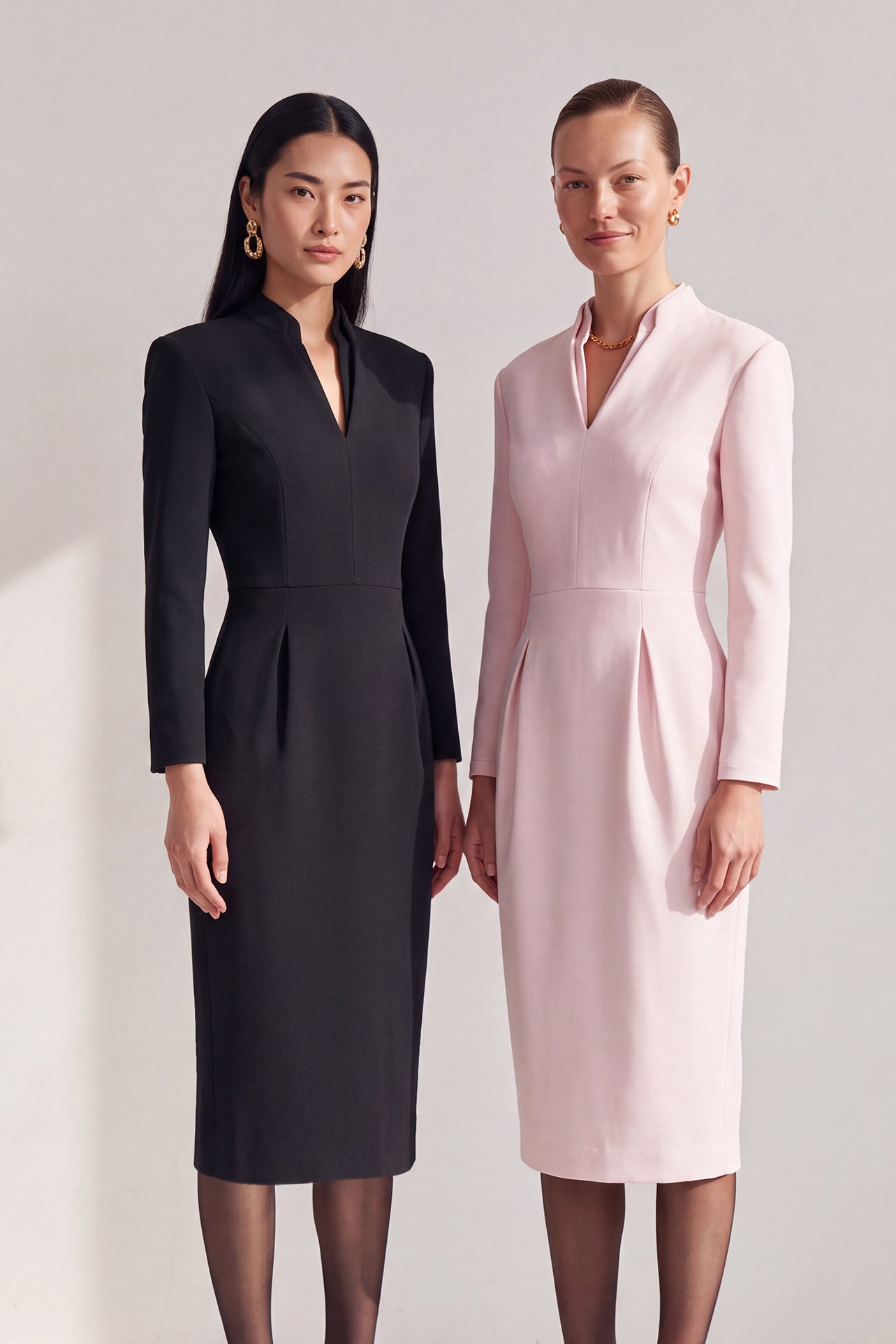 A style comparison of two elegant women modeling tailored long-sleeve midi dresses featuring a sophisticated stand collar with a split V-neckline and flattering double waist pleats; shown on the left in classic black and on the right in soft blush pink.