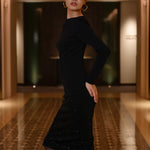 An elegant woman wearing a classic black long-sleeve sheath midi dress featuring a sophisticated boat neckline and a stunning gradient of sparkling rhinestones at the hemline for a refined evening look.