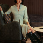 An elegant woman sitting on a modern chair wearing a sophisticated mint green textured suit featuring a cropped jacket with a mandarin collar, three white frog closures, and a matching high-waisted front-slit pencil skirt.