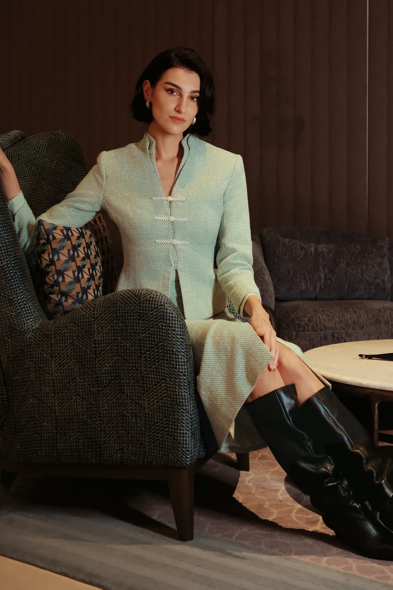 An elegant woman sitting on a modern chair wearing a sophisticated mint green textured suit featuring a cropped jacket with a mandarin collar, three white frog closures, and a matching high-waisted front-slit pencil skirt.
