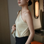 A stylish Asian woman modeling a sophisticated cream-colored asymmetric one-shoulder top featuring a single long sleeve, elegant diagonal draped pleats, and styled with high-waisted black trousers.