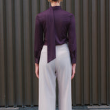 A chic woman wearing an elegant deep plum long-sleeve blouse with an asymmetric high-neck scarf tie, neatly tucked into a high-waisted black tweed skirt embellished with subtle sparkling sequins.