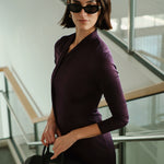 An elegant woman wearing a sophisticated deep plum-purple long-sleeve knit midi dress featuring a draped high-neck tie scarf and a dramatic asymmetrical flounce mermaid hemline.
