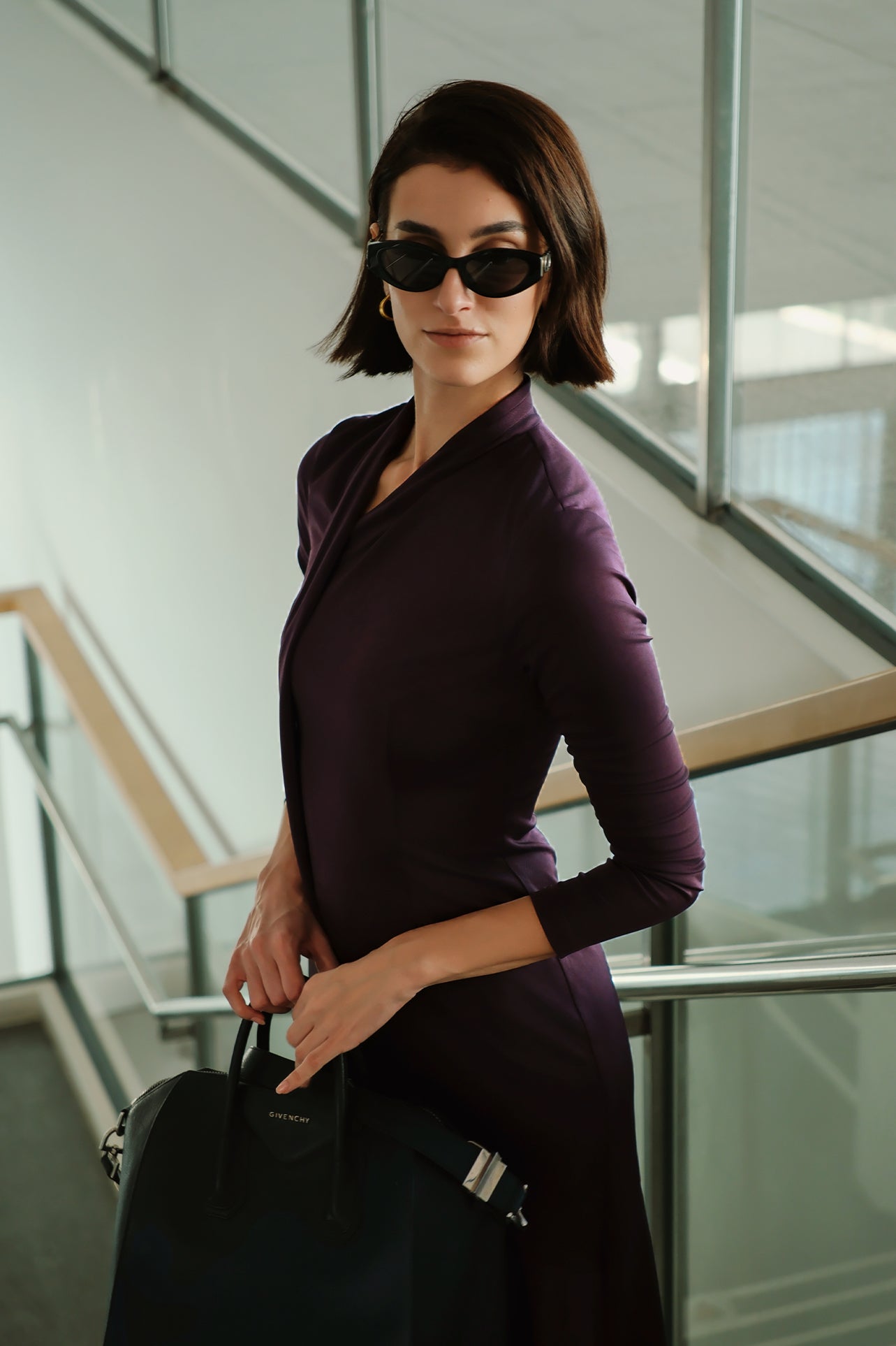 An elegant woman wearing a sophisticated deep plum-purple long-sleeve knit midi dress featuring a draped high-neck tie scarf and a dramatic asymmetrical flounce mermaid hemline.