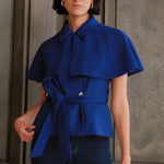 A style comparison of two professional women wearing modern cobalt blue outerwear featuring self-tie waist belts; on the left, a tailored cropped capelet jacket paired with jeans, and on the right, a double-breasted sleeveless vest with white contrast buttons styled with black trousers.