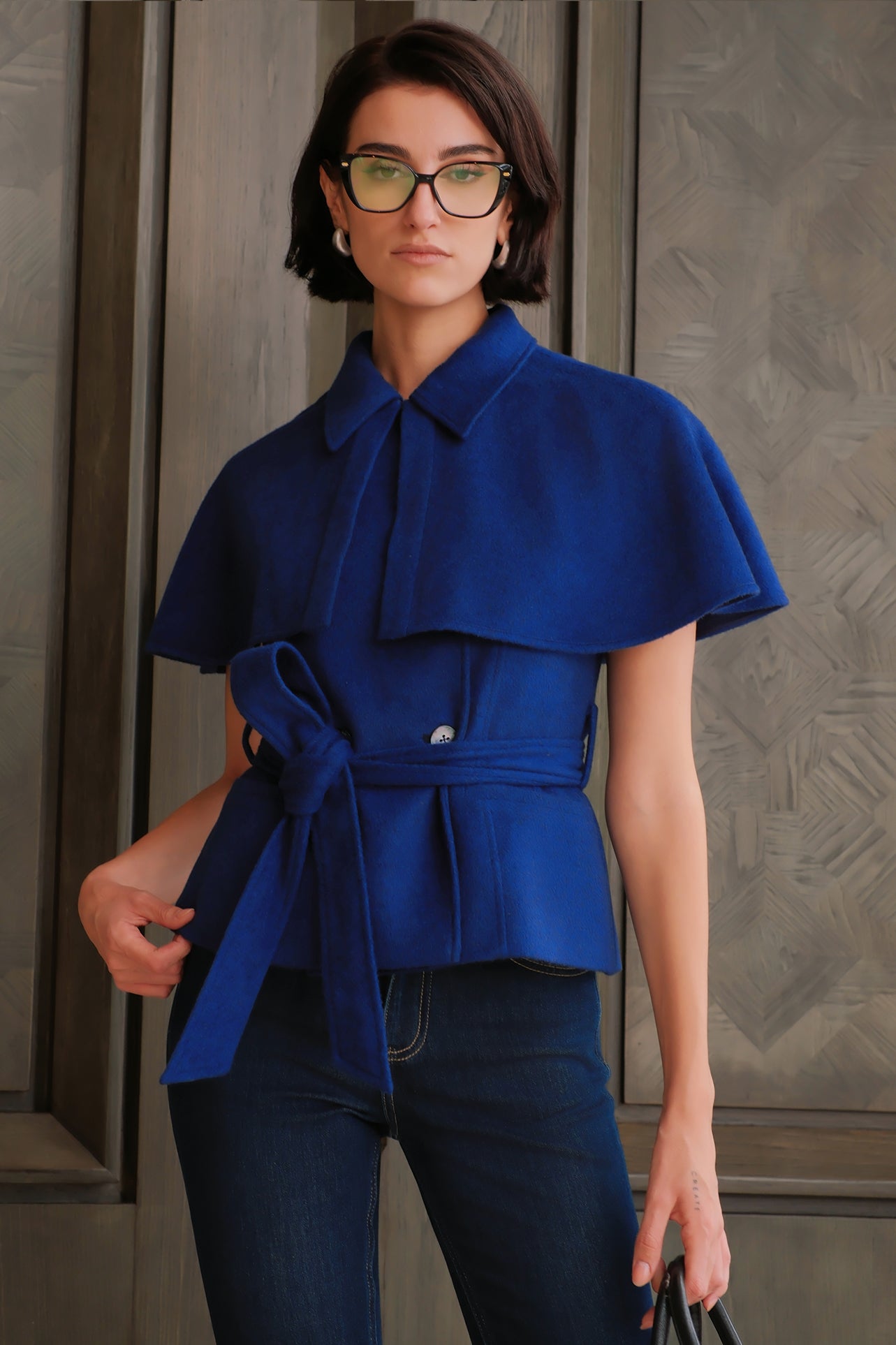 A style comparison of two professional women wearing modern cobalt blue outerwear featuring self-tie waist belts; on the left, a tailored cropped capelet jacket paired with jeans, and on the right, a double-breasted sleeveless vest with white contrast buttons styled with black trousers.