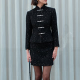 A chic woman wearing a luxurious black sparkle tweed tailored jacket featuring an authentic stand cheongsam collar, elegant horizontal pearl bar closures, and a flattering hourglass waistline.