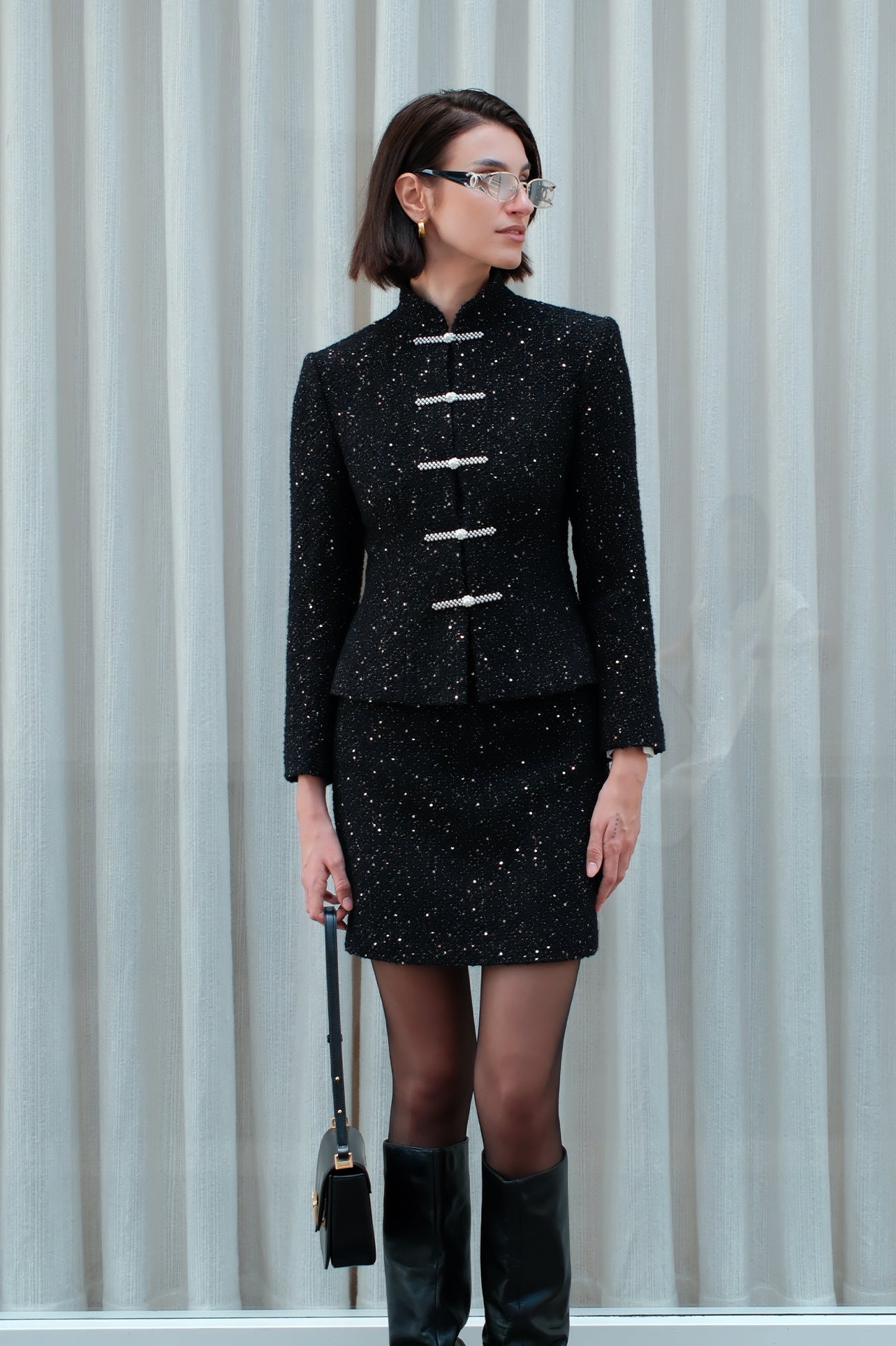 A chic woman wearing a luxurious black sparkle tweed tailored jacket featuring an authentic stand cheongsam collar, elegant horizontal pearl bar closures, and a flattering hourglass waistline.