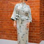 A professional woman modeling an elegant light sage green silk wrap jacket, featuring dark green bamboo leaf prints, a sophisticated mandarin collar with keyhole detail, and wide kimono sleeves.