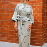 A professional woman modeling an elegant light sage green silk wrap jacket, featuring dark green bamboo leaf prints, a sophisticated mandarin collar with keyhole detail, and wide kimono sleeves.