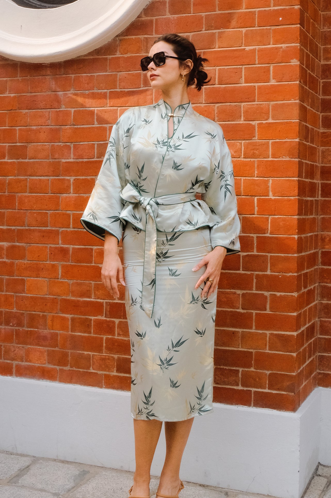 A professional woman modeling an elegant light sage green silk wrap jacket, featuring dark green bamboo leaf prints, a sophisticated mandarin collar with keyhole detail, and wide kimono sleeves.