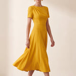 A blonde model wearing a sophisticated mustard yellow A-line midi dress featuring an elegant boat neckline with subtle shoulder pleating, short sleeves, and a flattering flared skirt.