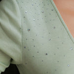 A style comparison of two elegant women's long-sleeve deep V-neck tops featuring subtle rhinestone crystal embellishments; shown on the left in nude camel tucked into tailored trousers, and on the right in pale mint green styled with a white skirt.