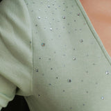 A style comparison of two elegant women's long-sleeve deep V-neck tops featuring subtle rhinestone crystal embellishments; shown on the left in nude camel tucked into tailored trousers, and on the right in pale mint green styled with a white skirt.