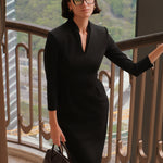 A style comparison of two elegant women modeling tailored long-sleeve midi dresses featuring a sophisticated stand collar with a split V-neckline and flattering double waist pleats; shown on the left in classic black and on the right in soft blush pink.