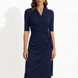 Two professional models wearing elegant wrap midi dresses with elbow-length sleeves; the left in a soft pastel lavender and the right in a deep navy blue, both featuring a sophisticated surplice V-neckline and slimming side ruched draping with a waist tie.