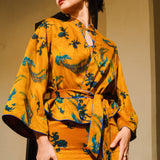 A beautiful woman modeling an elegant mustard yellow modern Chinese jacket, featuring intricate blue phoenix embroidery, a sophisticated mandarin collar, and wide kimono sleeves with a matching waist tie.