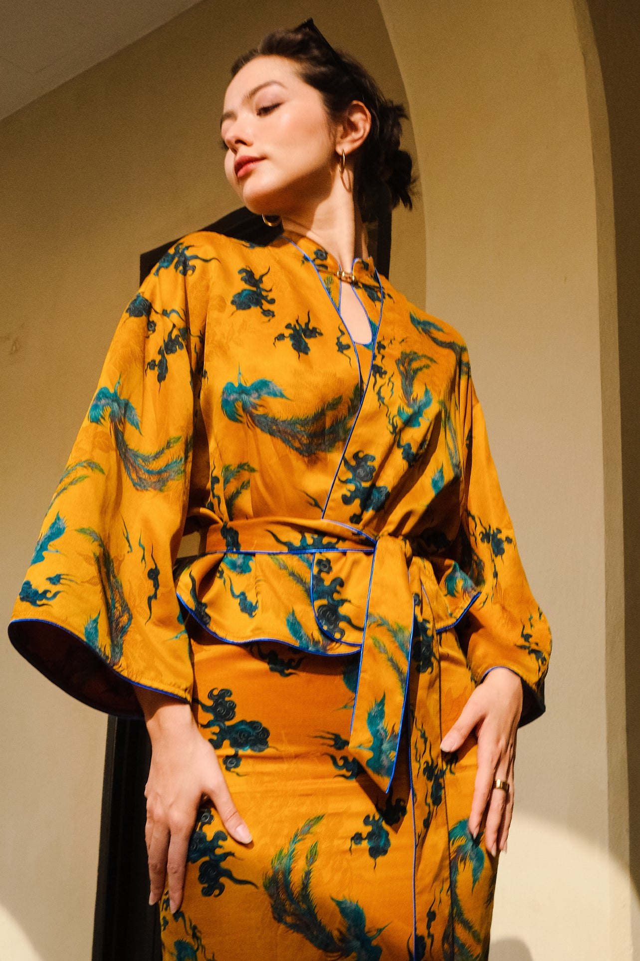 A beautiful woman modeling an elegant mustard yellow modern Chinese jacket, featuring intricate blue phoenix embroidery, a sophisticated mandarin collar, and wide kimono sleeves with a matching waist tie.