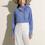 A style collection of three models presenting elegant dopamine office wear; the left in a white bow-tie blouse with a mustard yellow front-slit midi skirt, the center wearing a blue striped pussy-bow shirt with a straw hat and white wide-leg trousers, and the right in a vibrant orange satin blouse with a royal blue pencil skirt