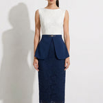 A professional model showcasing an elegant two-tone women's pantsuit, featuring a tailored cropped white blazer with a sharp deep V-neckline, paired with high-waisted navy blue trousers that have a unique curved overlay panel design and a golden metal hardware accent at the waist