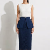 A professional model showcasing an elegant two-tone women's pantsuit, featuring a tailored cropped white blazer with a sharp deep V-neckline, paired with high-waisted navy blue trousers that have a unique curved overlay panel design and a golden metal hardware accent at the waist
