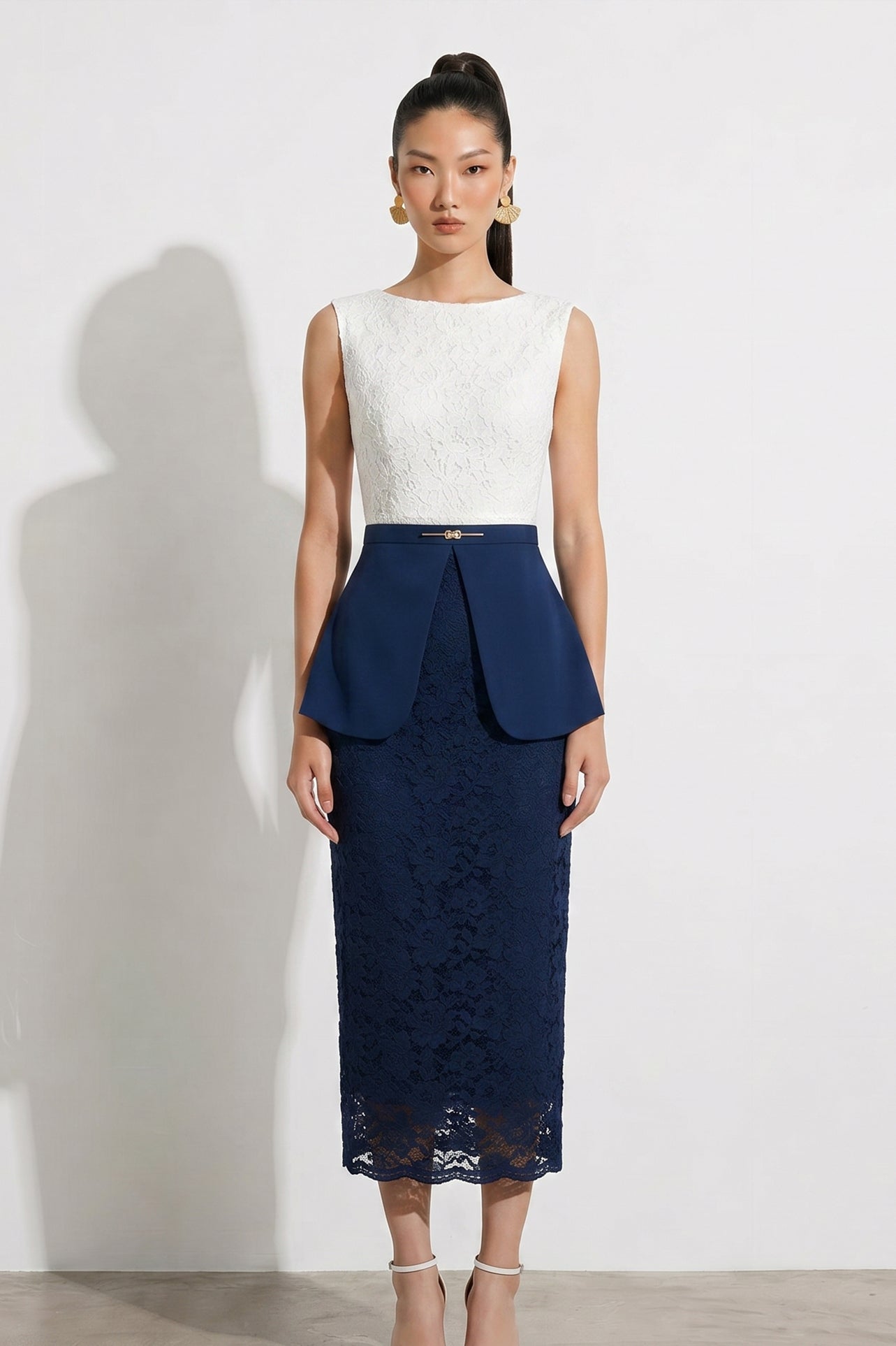 A professional model showcasing an elegant two-tone women's pantsuit, featuring a tailored cropped white blazer with a sharp deep V-neckline, paired with high-waisted navy blue trousers that have a unique curved overlay panel design and a golden metal hardware accent at the waist