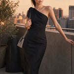 A style comparison of two elegant one-shoulder midi dresses; the left showcasing a sophisticated beige tailored sheath dress with a large tie-bow shoulder strap, and the right featuring the same design in timeless minimalist black.