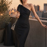 A style comparison of two elegant one-shoulder midi dresses; the left showcasing a sophisticated beige tailored sheath dress with a large tie-bow shoulder strap, and the right featuring the same design in timeless minimalist black.