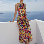 A beautiful professional model presenting an elegant tropical floral print halter maxi dress with a blue ocean background; featuring a sophisticated wrap tie-waist silhouette and vibrant multicolored botanical patterns for a chic summer resort look.