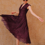 A chic woman wearing sunglasses and an elegant deep burgundy sleeveless V-neck A-line midi dress featuring textured jacquard fabric, delicate lace hemline, and a matching flowing neck scarf.