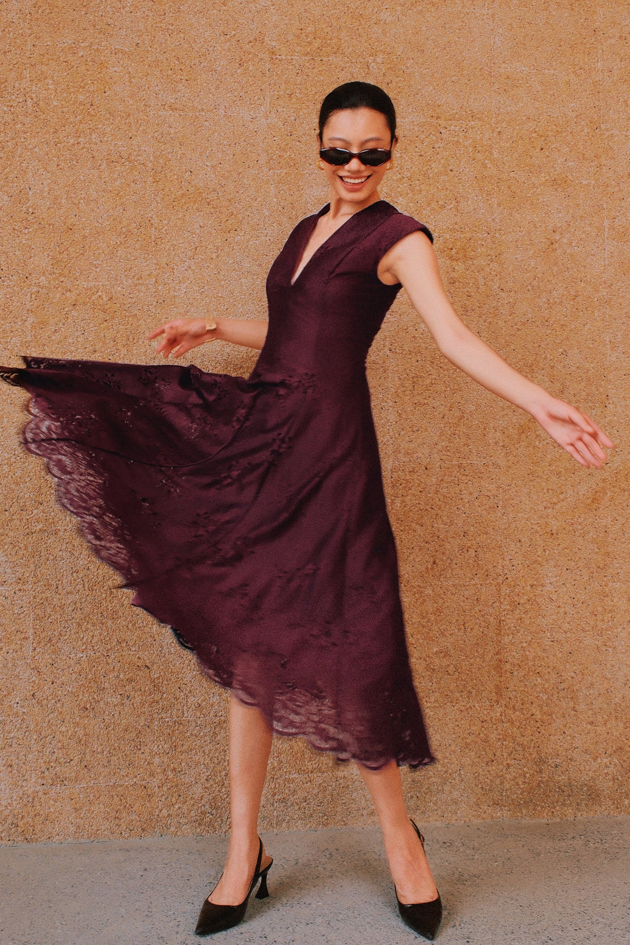 A chic woman wearing sunglasses and an elegant deep burgundy sleeveless V-neck A-line midi dress featuring textured jacquard fabric, delicate lace hemline, and a matching flowing neck scarf.