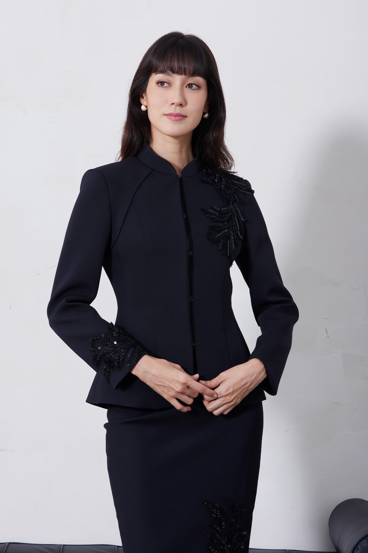 A professional woman wearing a sophisticated black skirt suit, featuring a mandarin collar with elegant leaf embroidery on the shoulder and skirt, and traditional frog button detailing for a modern oriental executive look.