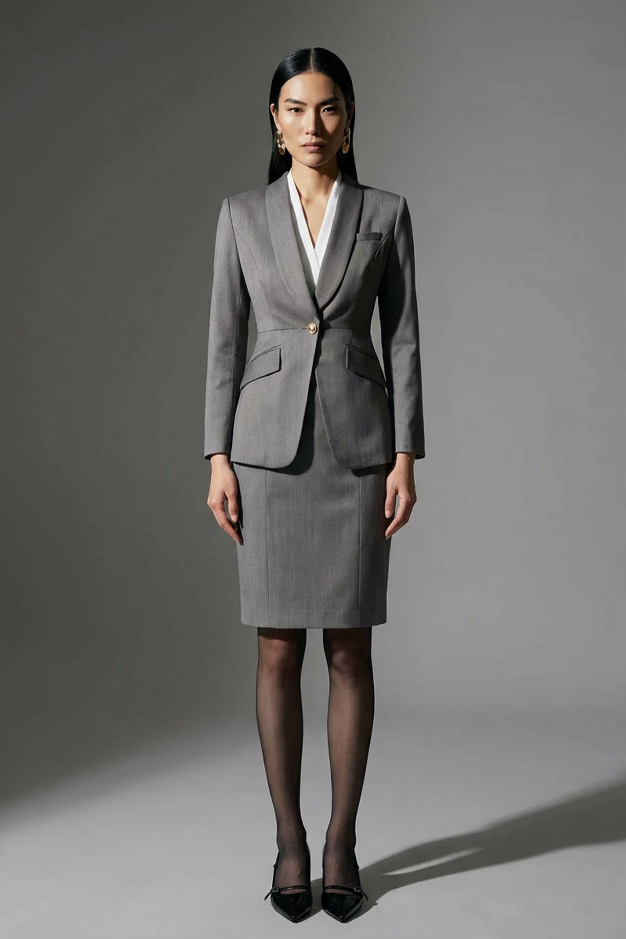 A professional woman wearing a sophisticated grey slim-fit blazer with a single gold button, featuring a tailored waist and notched lapels for an elegant executive office look.
