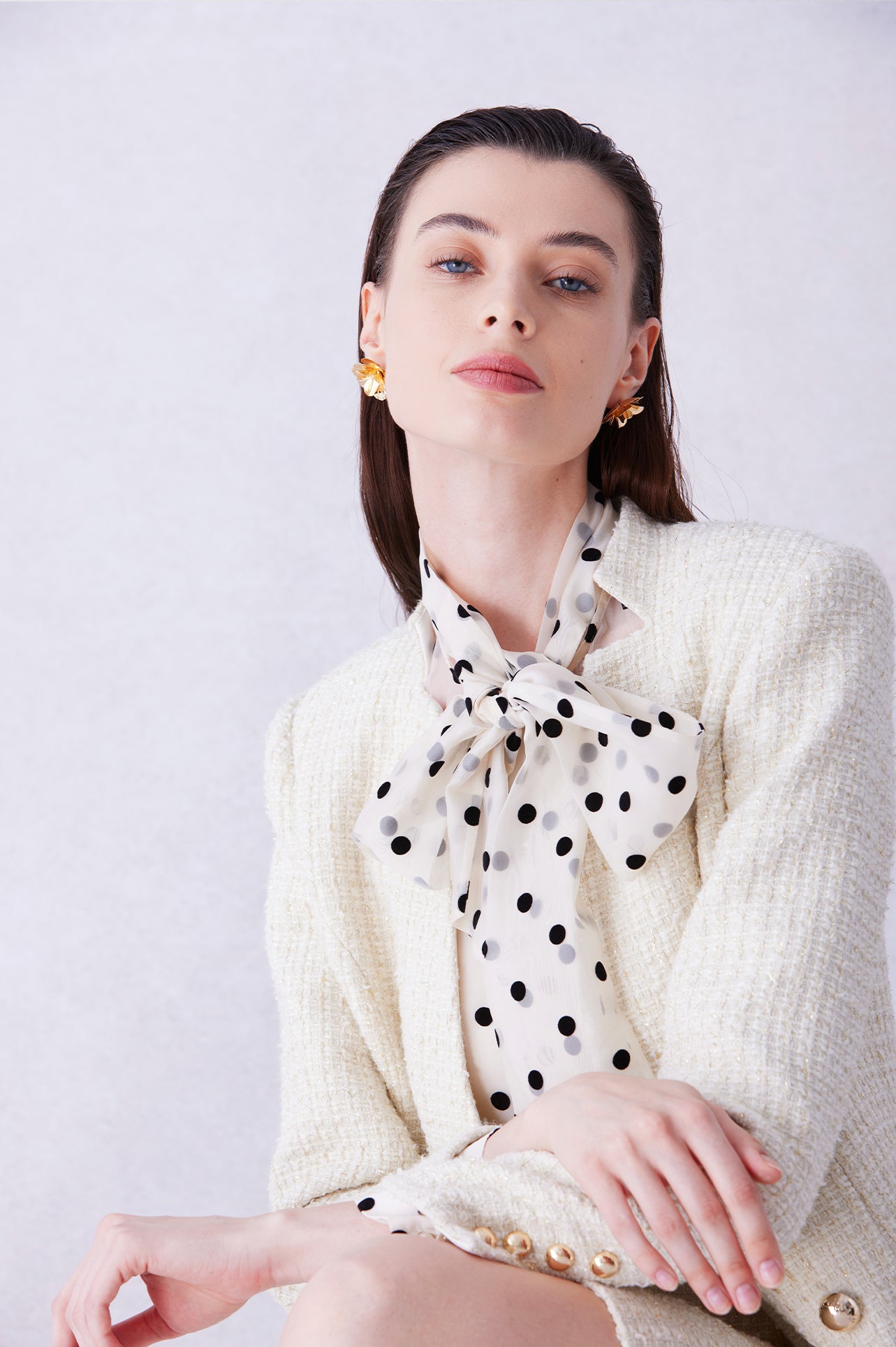 A professional woman wearing a sophisticated cream white tweed skirt suit, featuring a double-breasted blazer with gold buttons, a slim black belt, and an elegant polka dot pussy bow tie for a polished French chic office look.