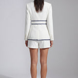 An elegant professional woman wearing a tailored white blazer with navy blue contrast piping, featuring a self-tie belt to define the waist for a smart-casual office look