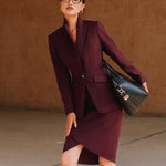 A professional woman wearing glasses and an elegant deep burgundy skirt suit featuring a sculptural stand-up lapel blazer with a single-button closure, tailored flap pockets, and a matching high-waisted pencil skirt.
