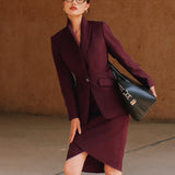 A professional woman wearing glasses and an elegant deep burgundy skirt suit featuring a sculptural stand-up lapel blazer with a single-button closure, tailored flap pockets, and a matching high-waisted pencil skirt.