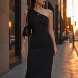 A style comparison of two elegant one-shoulder midi dresses; the left showcasing a sophisticated beige tailored sheath dress with a large tie-bow shoulder strap, and the right featuring the same design in timeless minimalist black.