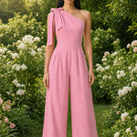 Two joyful women wearing elegant one-shoulder jumpsuits with large tie-bow shoulder straps in a sunny park; one jumps in a pastel pink wide-leg jumpsuit with yellow sneakers, while the other claps in a beige ankle-length jumpsuit.