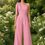 Two joyful women wearing elegant one-shoulder jumpsuits with large tie-bow shoulder straps in a sunny park; one jumps in a pastel pink wide-leg jumpsuit with yellow sneakers, while the other claps in a beige ankle-length jumpsuit.
