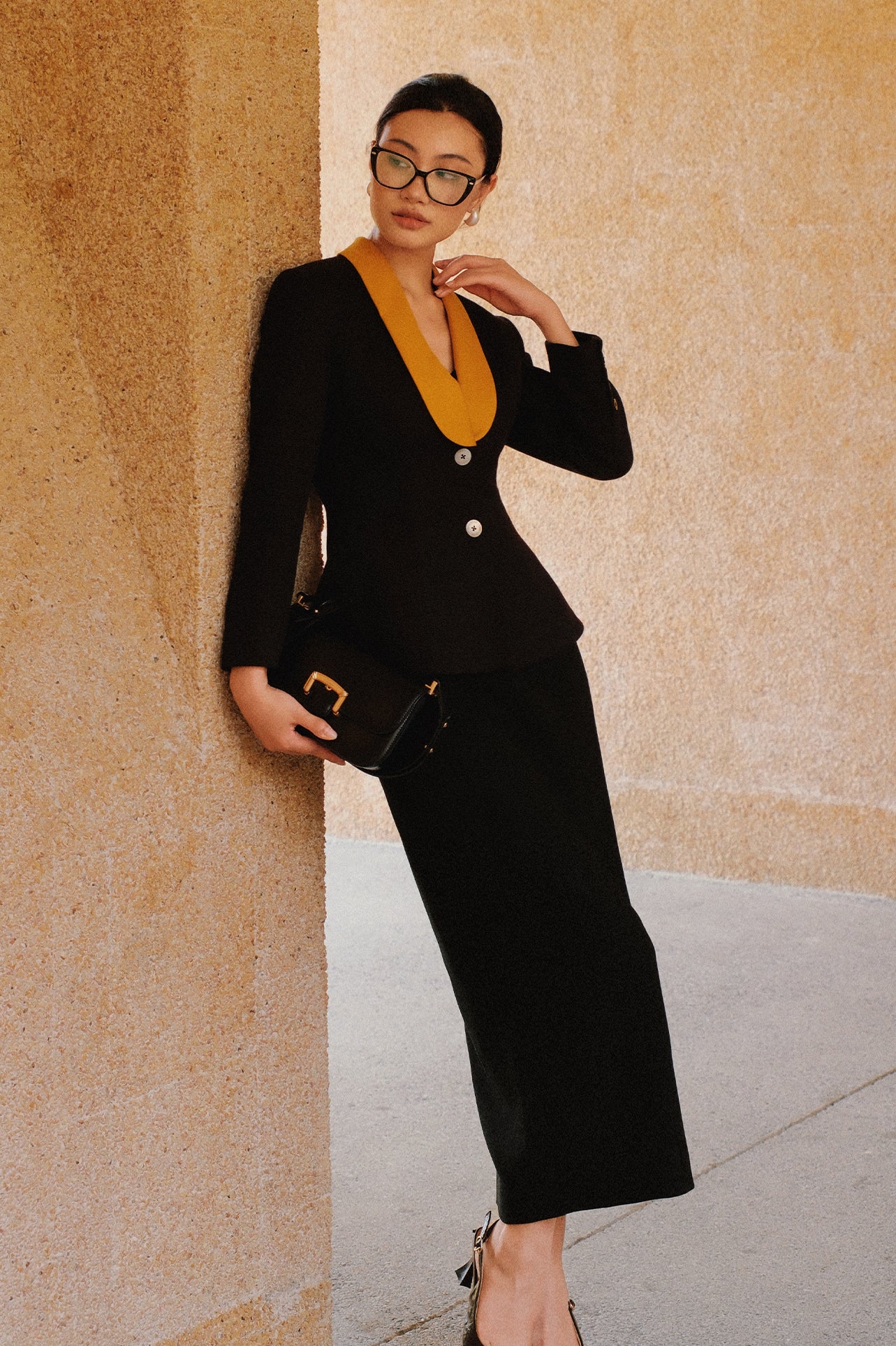 A professional Asian woman wearing glasses and an elegant black tailored blazer featuring a striking mustard yellow contrast shawl lapel, statement front button, and buttoned cuffs for a sophisticated intellectual corporate look.